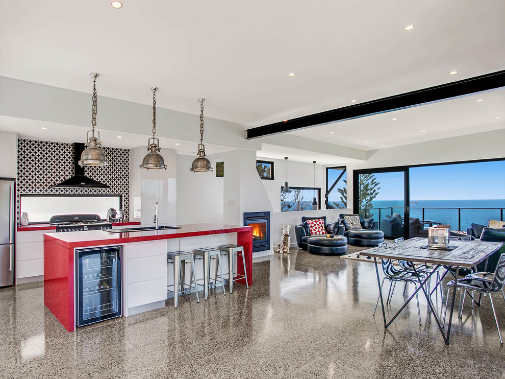 This is an example of a galley open plan kitchen in Wollongong with an integrated sink, flat-panel cabinets, white cabinets, composite countertops, black splashback, stainless steel appliances, concrete flooring, an island, grey floors and red worktops.