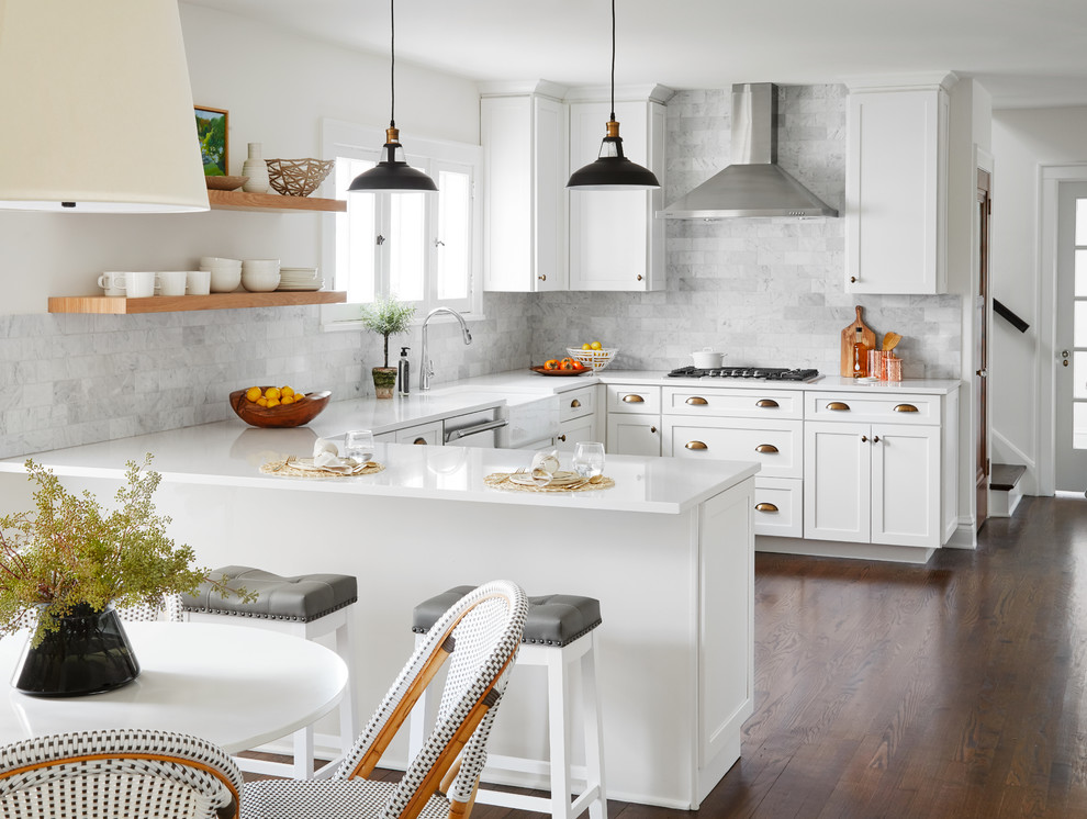 This is an example of a medium sized traditional u-shaped kitchen/diner in Chicago with a belfast sink, shaker cabinets, white cabinets, engineered stone countertops, grey splashback, marble splashback, stainless steel appliances, medium hardwood flooring, no island, brown floors and white worktops.
