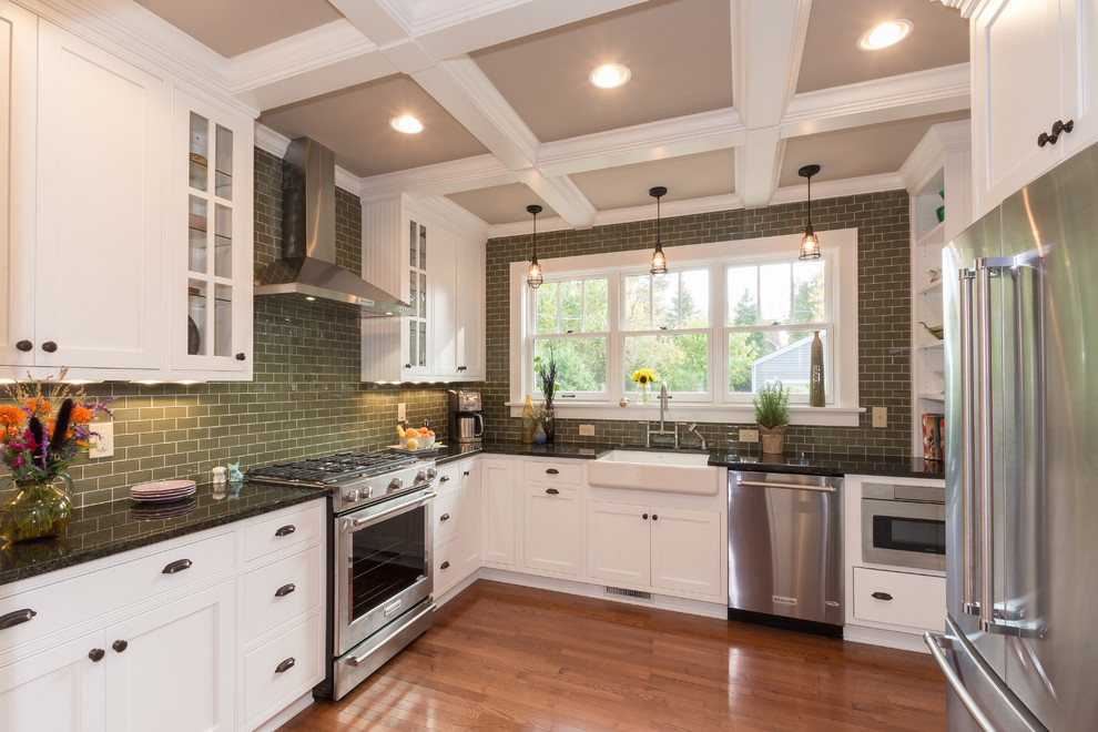 Example of a mid-sized transitional l-shaped medium tone wood floor enclosed kitchen design in Columbus with a farmhouse sink, beaded inset cabinets, white cabinets, granite countertops, green backsplash, glass tile backsplash, stainless steel appliances and no island