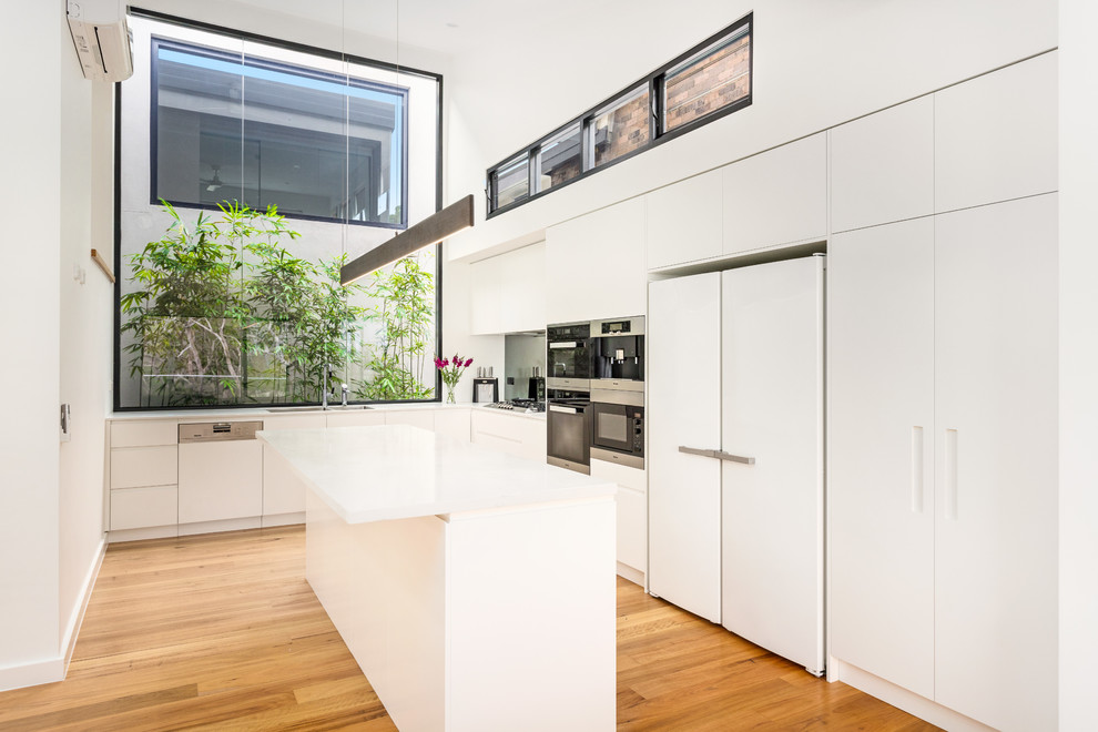 Mittelgroße Moderne Küche in L-Form mit Einbauwaschbecken, flächenbündigen Schrankfronten, weißen Schränken, Küchengeräten aus Edelstahl, braunem Holzboden, zwei Kücheninseln, braunem Boden, weißer Arbeitsplatte, Küchenrückwand in Grau und Glasrückwand in Sydney