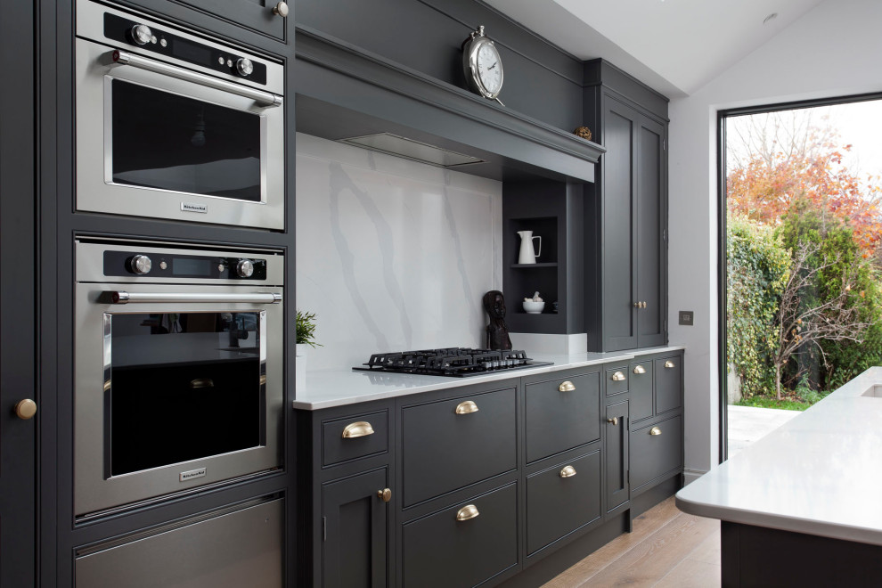 Medium sized traditional single-wall open plan kitchen in Dublin with a single-bowl sink, shaker cabinets, grey cabinets, quartz worktops, white splashback, engineered quartz splashback, stainless steel appliances, light hardwood flooring, an island, brown floors, white worktops and a vaulted ceiling.