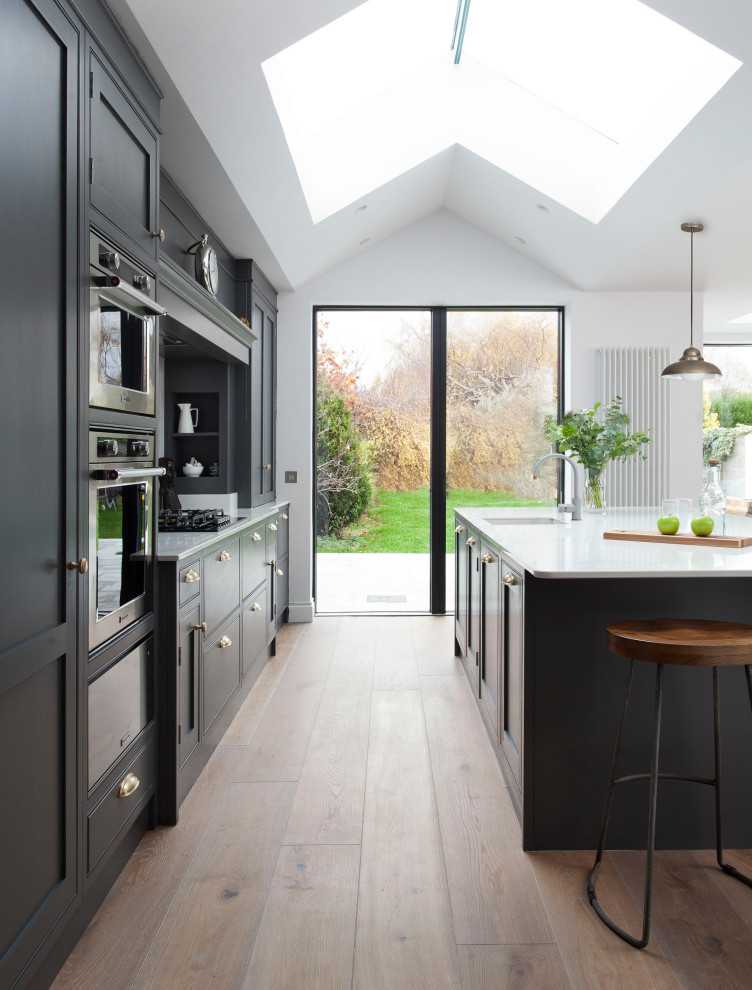 This is an example of a medium sized classic single-wall open plan kitchen in Dublin with a single-bowl sink, shaker cabinets, grey cabinets, quartz worktops, white splashback, engineered quartz splashback, stainless steel appliances, light hardwood flooring, an island, brown floors, white worktops and a vaulted ceiling.