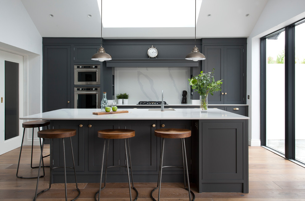 This is an example of a large traditional galley kitchen in Dublin with a single-bowl sink, shaker cabinets, grey cabinets, quartz worktops, white splashback, stone slab splashback, stainless steel appliances, medium hardwood flooring, an island, brown floors and white worktops.