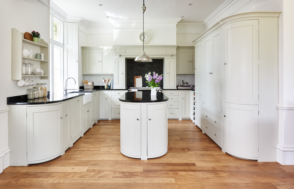Classic u-shaped kitchen in Wiltshire with a belfast sink, shaker cabinets, beige cabinets, black splashback, light hardwood flooring, an island and black worktops.