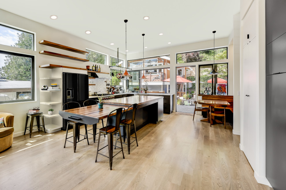Design ideas for a large contemporary l-shaped open plan kitchen in Seattle with a belfast sink, flat-panel cabinets, grey cabinets, wood worktops, white splashback, metro tiled splashback, black appliances, light hardwood flooring, an island and white worktops.