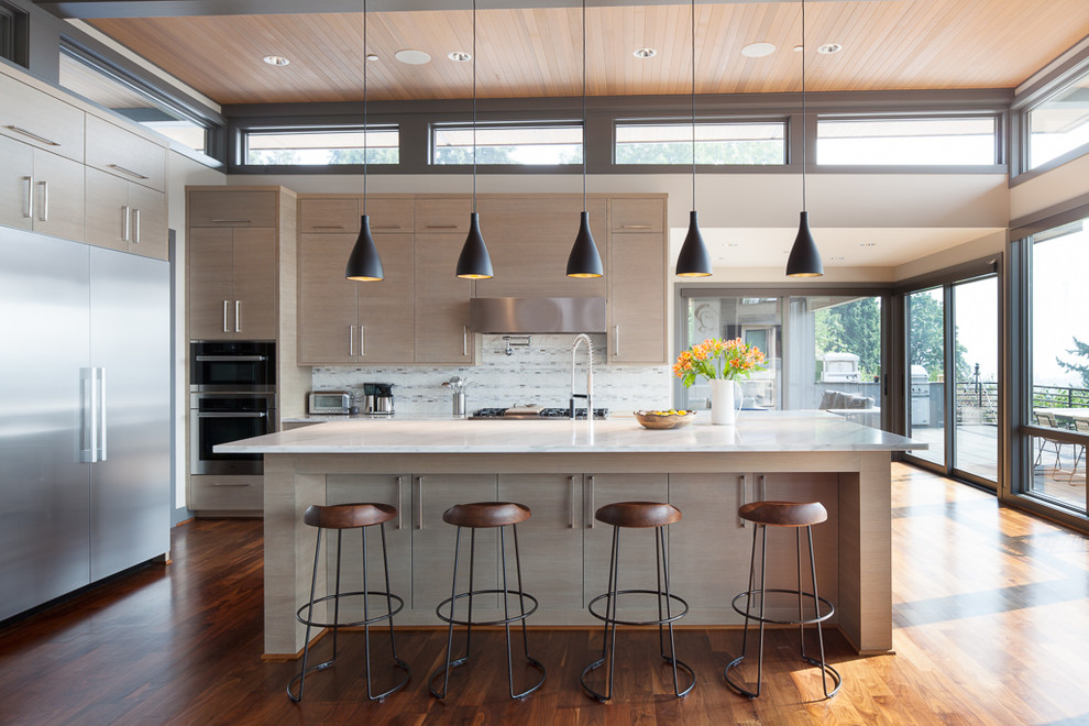 Modern inredning av ett kök, med släta luckor, skåp i ljust trä, flerfärgad stänkskydd, rostfria vitvaror, mörkt trägolv, en köksö och brunt golv