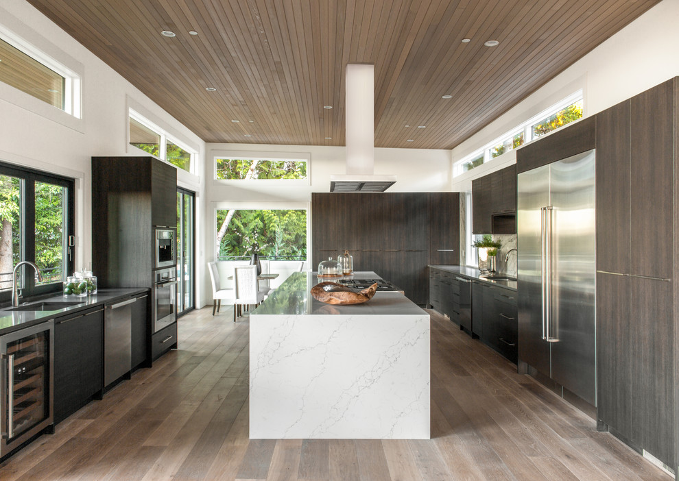 Cette image montre une cuisine design en U et bois foncé avec un évier encastré, un placard à porte plane, un électroménager en acier inoxydable, un sol en bois brun, îlot, un sol marron et un plan de travail blanc.