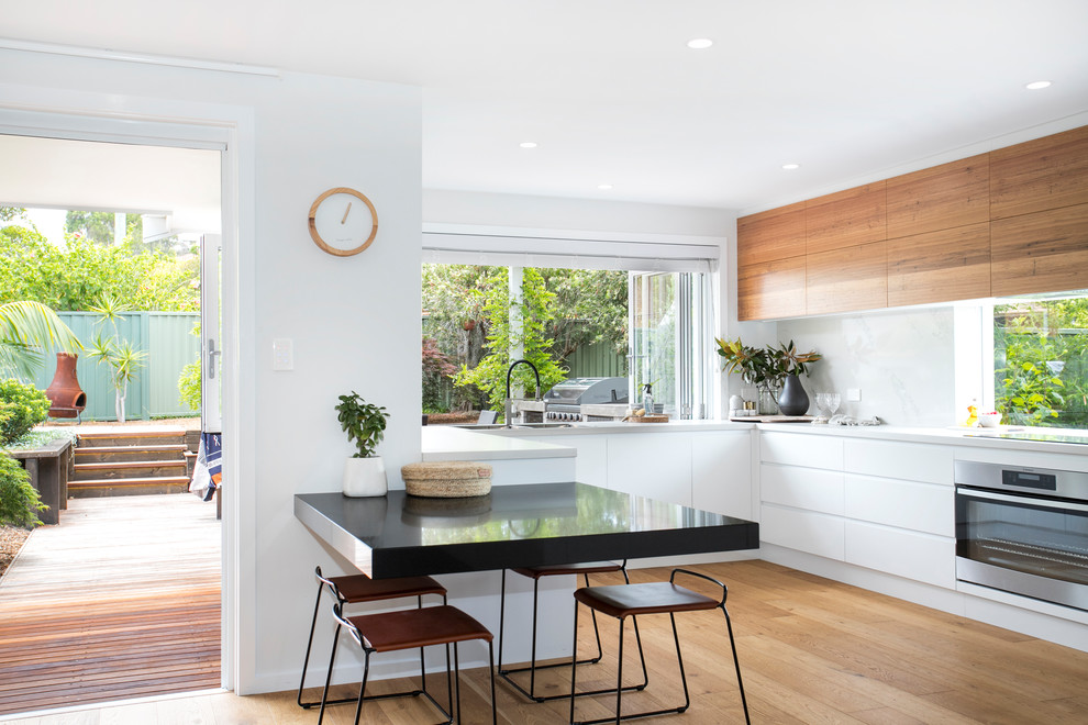 Example of a trendy u-shaped medium tone wood floor and brown floor eat-in kitchen design in Wollongong with an undermount sink, flat-panel cabinets, white cabinets, white backsplash, stainless steel appliances and white countertops