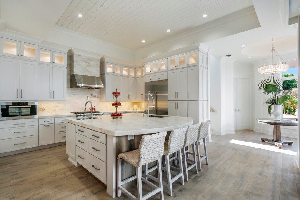 Photo of a large coastal l-shaped kitchen/diner in Miami with a submerged sink, shaker cabinets, white cabinets, marble worktops, white splashback, porcelain splashback, stainless steel appliances, light hardwood flooring and an island.