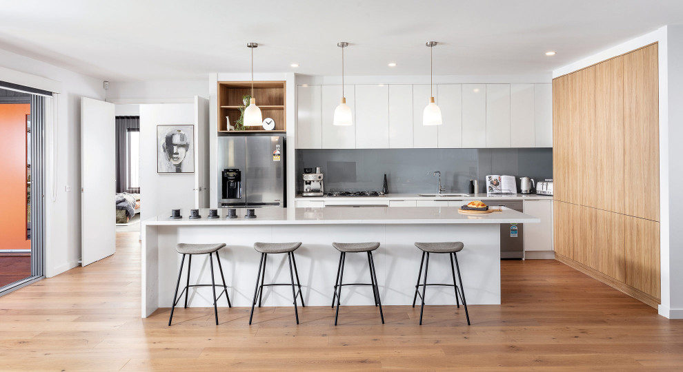 Moderne Küche in L-Form mit Unterbauwaschbecken, flächenbündigen Schrankfronten, weißen Schränken, Küchenrückwand in Grau, Küchengeräten aus Edelstahl, braunem Holzboden, Kücheninsel, braunem Boden und weißer Arbeitsplatte in Melbourne