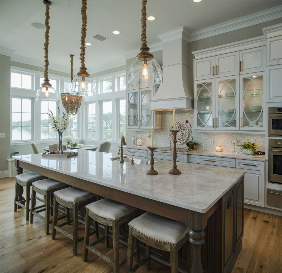 Beach style medium tone wood floor and brown floor eat-in kitchen photo in Tampa with a farmhouse sink, raised-panel cabinets, white cabinets, beige backsplash, mosaic tile backsplash, stainless steel appliances, an island and beige countertops