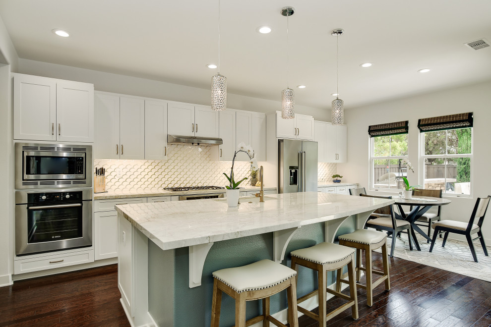 Photo of a large classic galley open plan kitchen in San Diego with a single-bowl sink, shaker cabinets, white cabinets, quartz worktops, white splashback, glass tiled splashback, stainless steel appliances, dark hardwood flooring, an island, brown floors and white worktops.