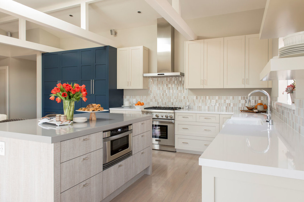 Example of a beach style l-shaped light wood floor kitchen design in San Francisco with a farmhouse sink, shaker cabinets, beige cabinets, beige backsplash, stainless steel appliances and an island