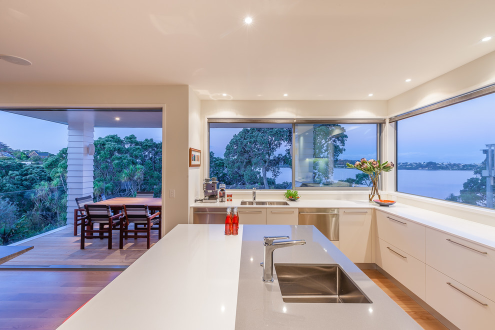 Example of a mid-sized island style l-shaped light wood floor kitchen pantry design in Auckland with an undermount sink, flat-panel cabinets, white cabinets, quartz countertops, green backsplash, glass sheet backsplash, stainless steel appliances and an island