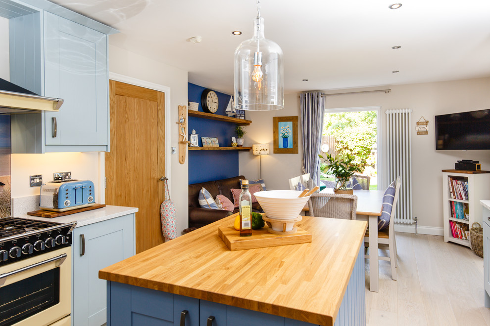 Beach style u-shaped open plan kitchen in Cambridgeshire with a belfast sink, shaker cabinets, blue cabinets, wood worktops, glass sheet splashback, integrated appliances, light hardwood flooring and an island.