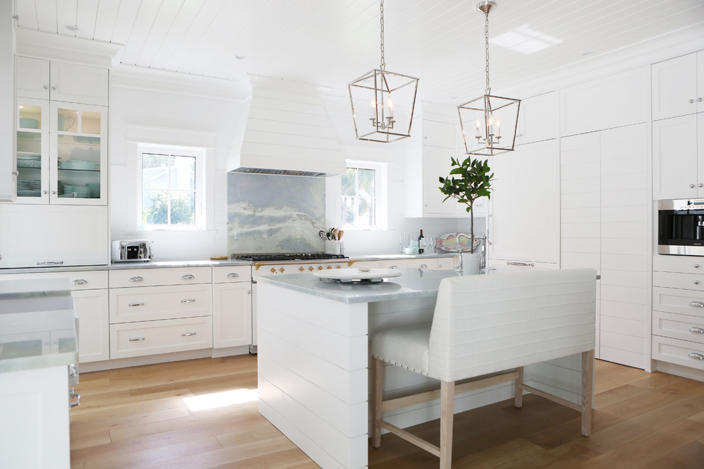 This is an example of a beach style kitchen in Miami with shaker cabinets, white cabinets, multi-coloured splashback, stone slab splashback, white appliances, light hardwood flooring, an island, beige floors and grey worktops.