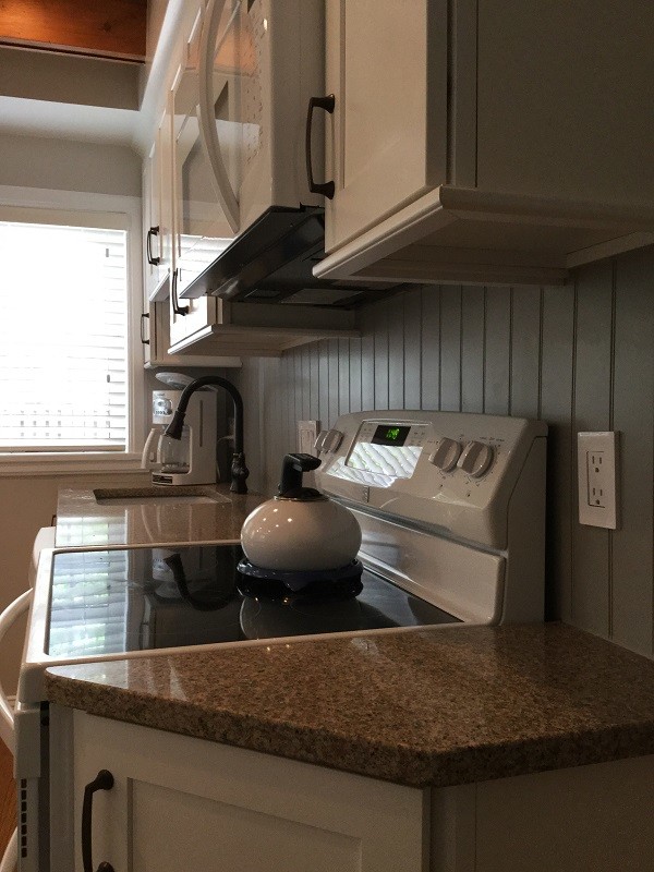 Photo of a medium sized coastal single-wall kitchen/diner in DC Metro with a submerged sink, shaker cabinets, white cabinets, engineered stone countertops, grey splashback, wood splashback, white appliances, medium hardwood flooring, an island, brown floors and brown worktops.