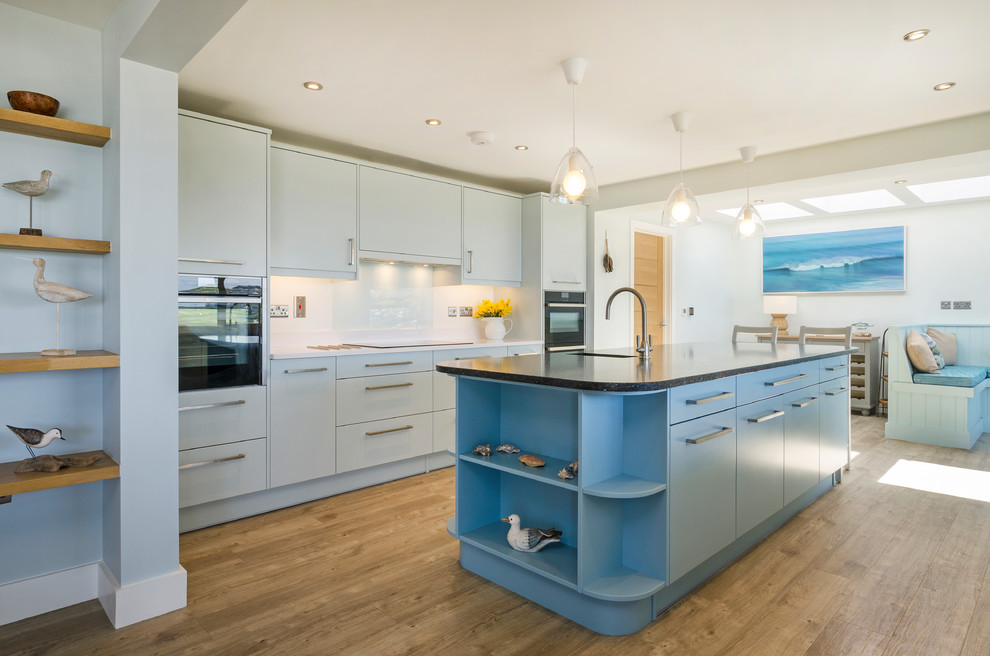 Design ideas for a nautical open plan kitchen in Cornwall with a single-bowl sink, flat-panel cabinets, blue cabinets, white splashback, glass sheet splashback, black appliances, light hardwood flooring and an island.