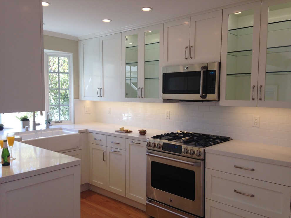 This is an example of a traditional u-shaped kitchen in Los Angeles with a belfast sink, shaker cabinets, white cabinets, engineered stone countertops, white splashback, metro tiled splashback, integrated appliances and medium hardwood flooring.