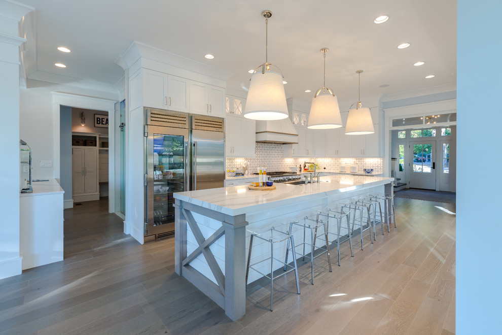 Example of a large l-shaped medium tone wood floor and gray floor kitchen pantry design in Other with a farmhouse sink, shaker cabinets, white cabinets, quartz countertops, white backsplash, marble backsplash, stainless steel appliances, an island and white countertops