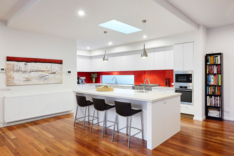 Foto på ett mellanstort funkis kök, med en dubbel diskho, luckor med infälld panel, vita skåp, granitbänkskiva, rött stänkskydd, glaspanel som stänkskydd, rostfria vitvaror, mellanmörkt trägolv och en köksö
