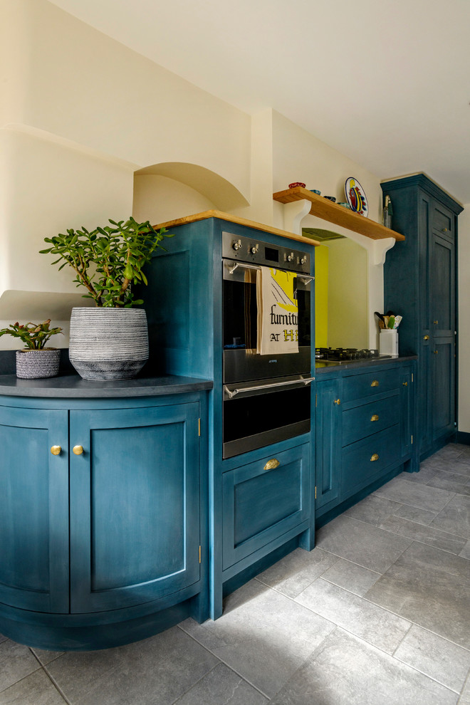 This is an example of a classic kitchen in London with a belfast sink, shaker cabinets, blue cabinets, granite worktops, yellow splashback, glass sheet splashback, ceramic flooring and black worktops.