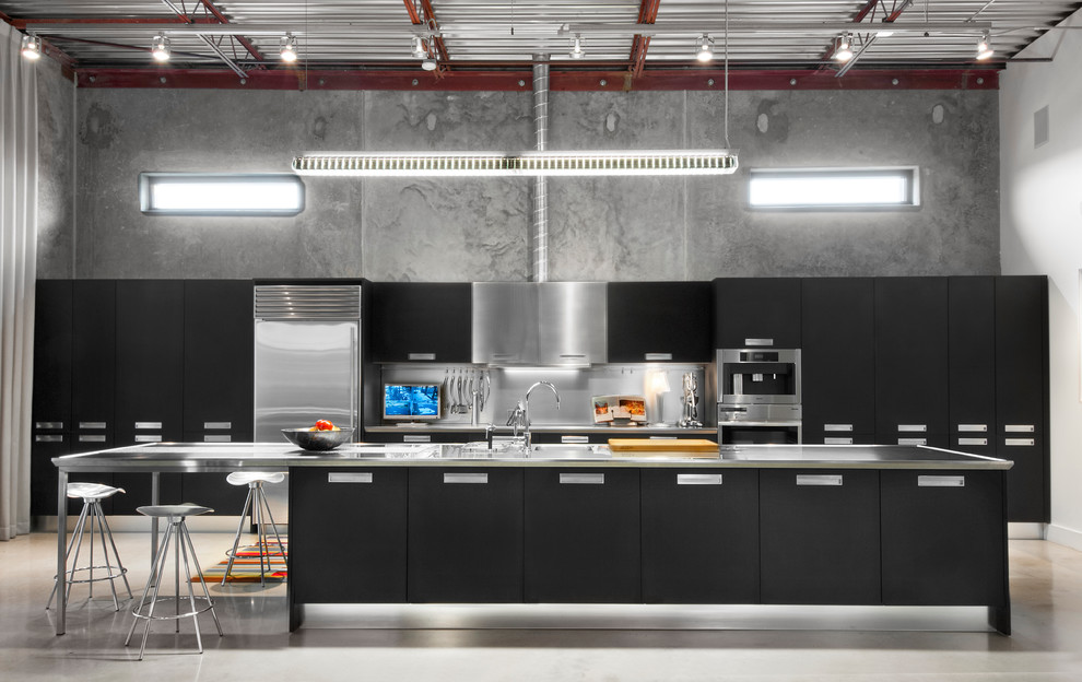 Foto på ett industriellt kök, med en integrerad diskho, släta luckor, svarta skåp, bänkskiva i rostfritt stål, stänkskydd med metallisk yta, rostfria vitvaror, betonggolv och en köksö