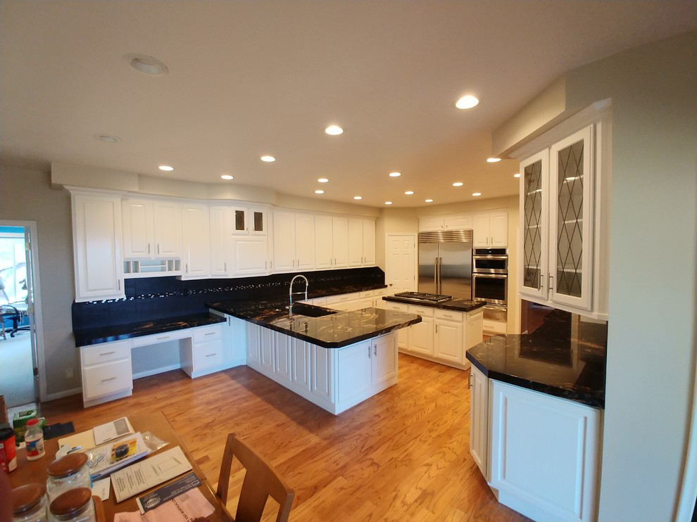 Bild på ett stort eklektiskt svart svart kök, med en undermonterad diskho, luckor med upphöjd panel, vita skåp, granitbänkskiva, svart stänkskydd, stänkskydd i porslinskakel, rostfria vitvaror, ljust trägolv, en köksö och beiget golv