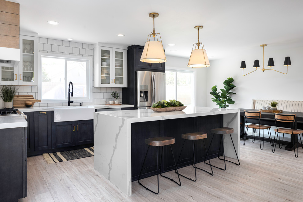 Example of a transitional light wood floor and beige floor eat-in kitchen design in Orange County with a farmhouse sink, shaker cabinets, black cabinets, white backsplash, stainless steel appliances, an island and gray countertops