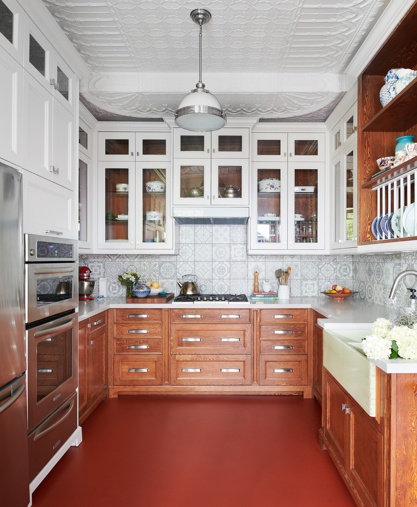 Mid-sized eclectic u-shaped red floor and concrete floor enclosed kitchen photo in Toronto with a farmhouse sink, shaker cabinets, medium tone wood cabinets, gray backsplash, ceramic backsplash, stainless steel appliances, no island, white countertops and solid surface countertops