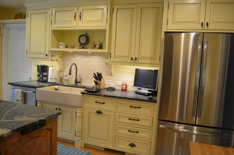 Example of a mid-sized country u-shaped light wood floor eat-in kitchen design in Other with a farmhouse sink, glass-front cabinets, yellow cabinets, soapstone countertops, white backsplash, glass tile backsplash, stainless steel appliances and an island