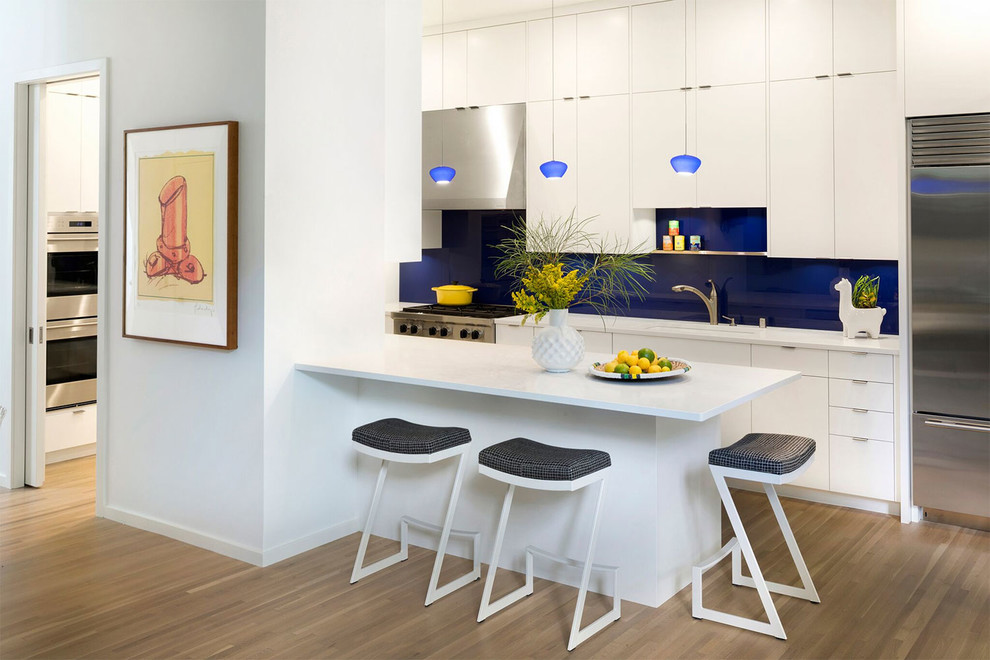 Contemporary l-shaped kitchen in Minneapolis with a submerged sink, flat-panel cabinets, white cabinets, blue splashback, glass sheet splashback, stainless steel appliances, medium hardwood flooring, a breakfast bar and brown floors.