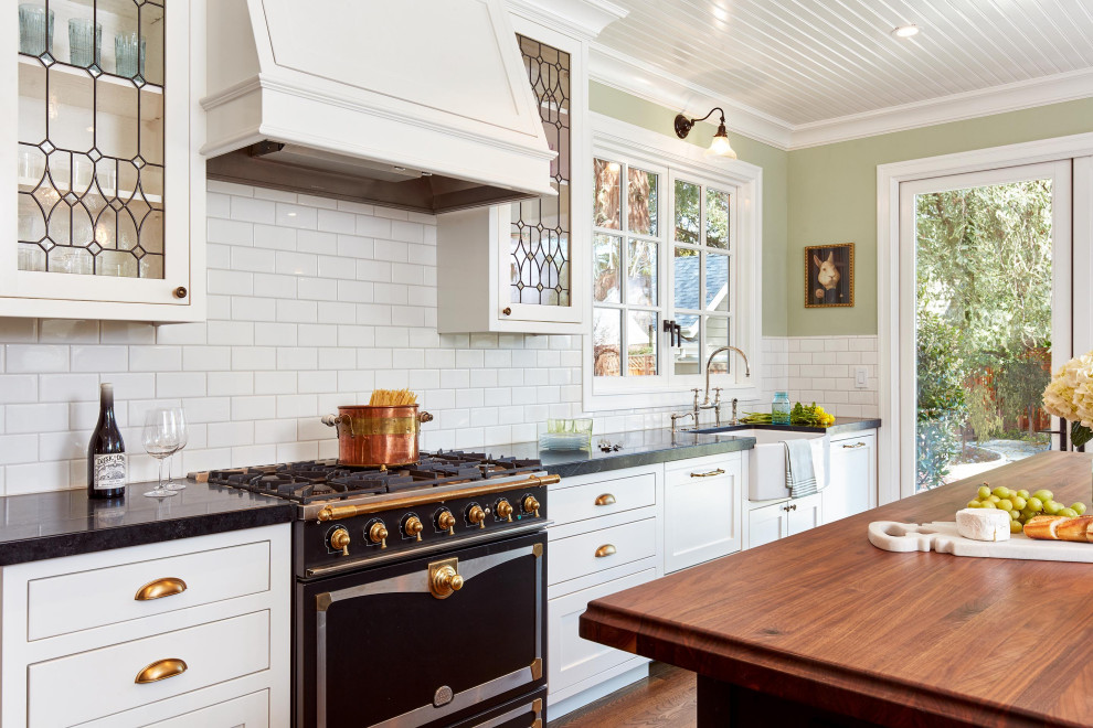 Enclosed kitchen - large traditional galley medium tone wood floor, brown floor and shiplap ceiling enclosed kitchen idea in San Francisco with a farmhouse sink, shaker cabinets, white cabinets, white backsplash, subway tile backsplash, black appliances, an island and black countertops