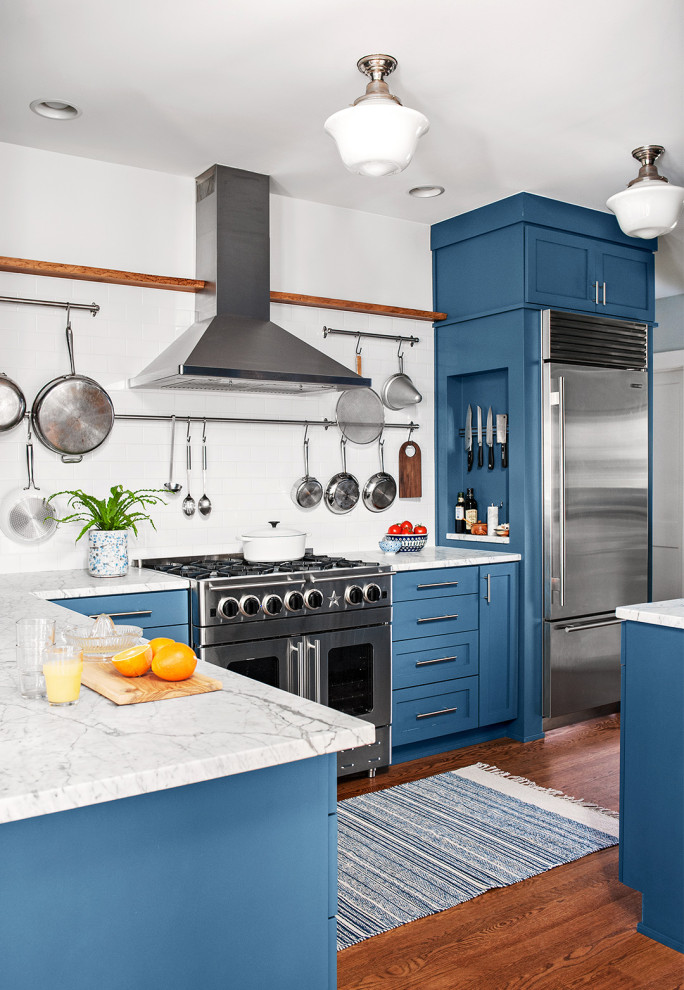Imagen de cocina tradicional renovada con armarios estilo shaker, puertas de armario azules, electrodomésticos de acero inoxidable, suelo de madera en tonos medios, península y encimeras blancas