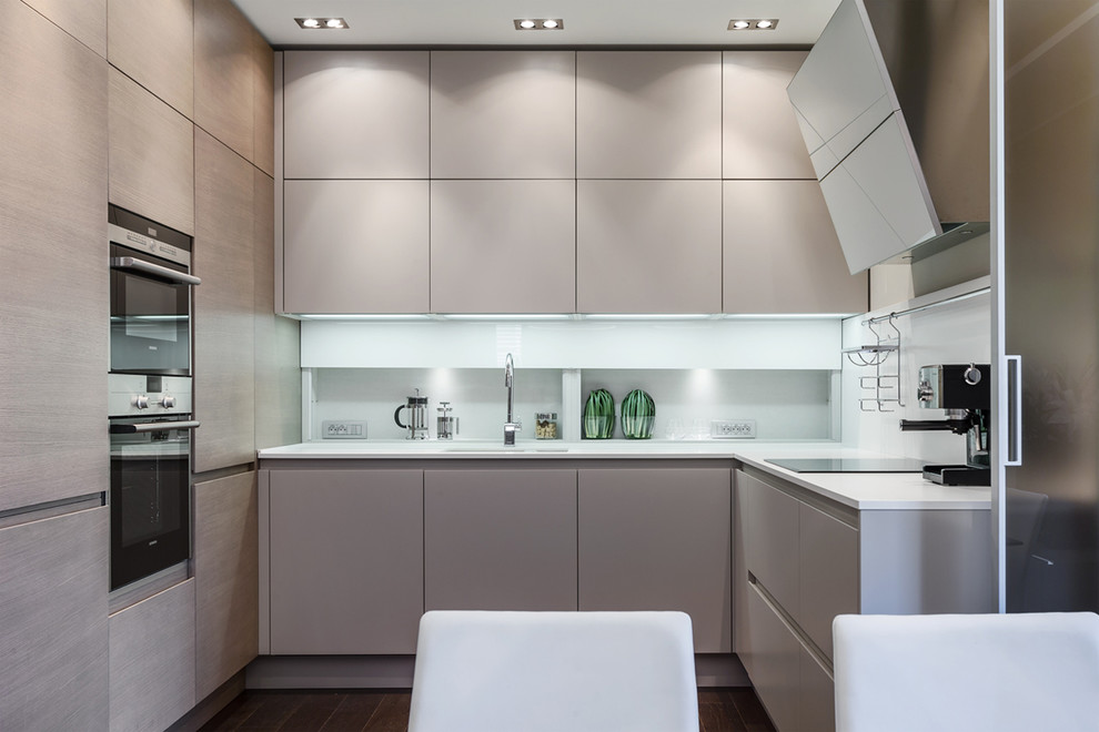 Moderne Wohnküche in L-Form mit flächenbündigen Schrankfronten, beigen Schränken, Küchengeräten aus Edelstahl, Mineralwerkstoff-Arbeitsplatte und Küchenrückwand in Weiß in Sonstige
