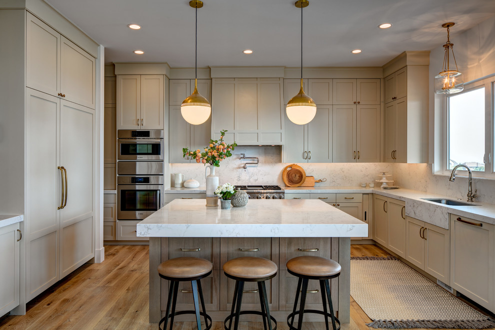 Inspiration for a large traditional u-shaped kitchen in Salt Lake City with recessed-panel cabinets, marble worktops, marble splashback, stainless steel appliances, an island, a submerged sink, beige cabinets, white splashback, medium hardwood flooring, brown floors and white worktops.