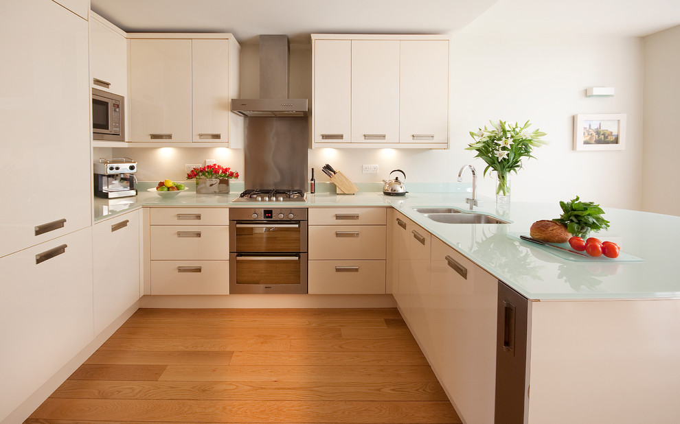 This is an example of a small contemporary u-shaped kitchen in London with a double-bowl sink, flat-panel cabinets, white cabinets, glass worktops, stainless steel appliances, medium hardwood flooring, a breakfast bar and turquoise worktops.