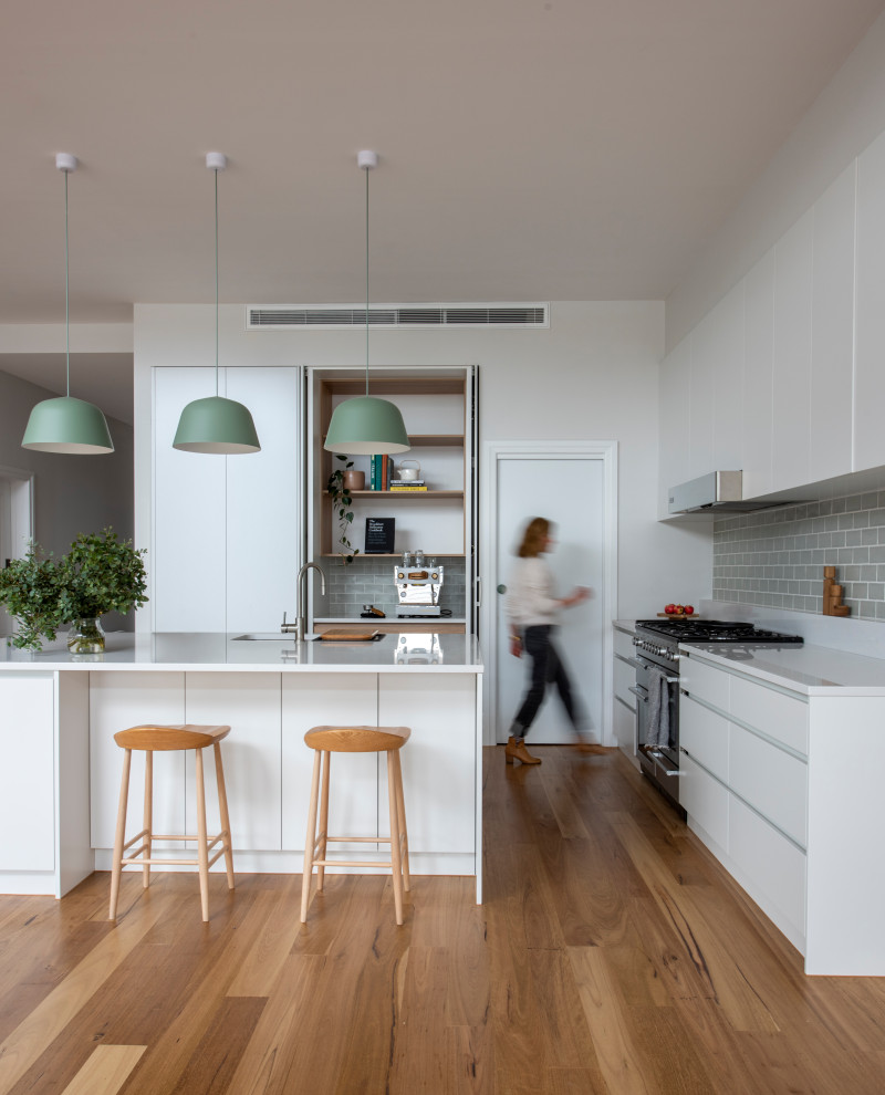 Inspiration for a large contemporary l-shaped kitchen/diner in Melbourne with a double-bowl sink, flat-panel cabinets, white cabinets, engineered stone countertops, green splashback, metro tiled splashback, stainless steel appliances, medium hardwood flooring, an island, brown floors and white worktops.
