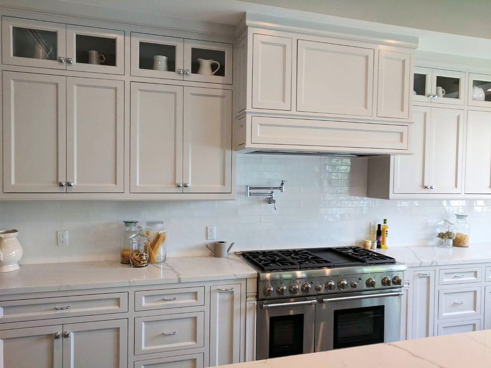 Modern inredning av ett stort vit vitt kök, med en integrerad diskho, vita skåp, marmorbänkskiva, vitt stänkskydd, stänkskydd i porslinskakel, rostfria vitvaror, ljust trägolv, en köksö, grått golv och luckor med glaspanel