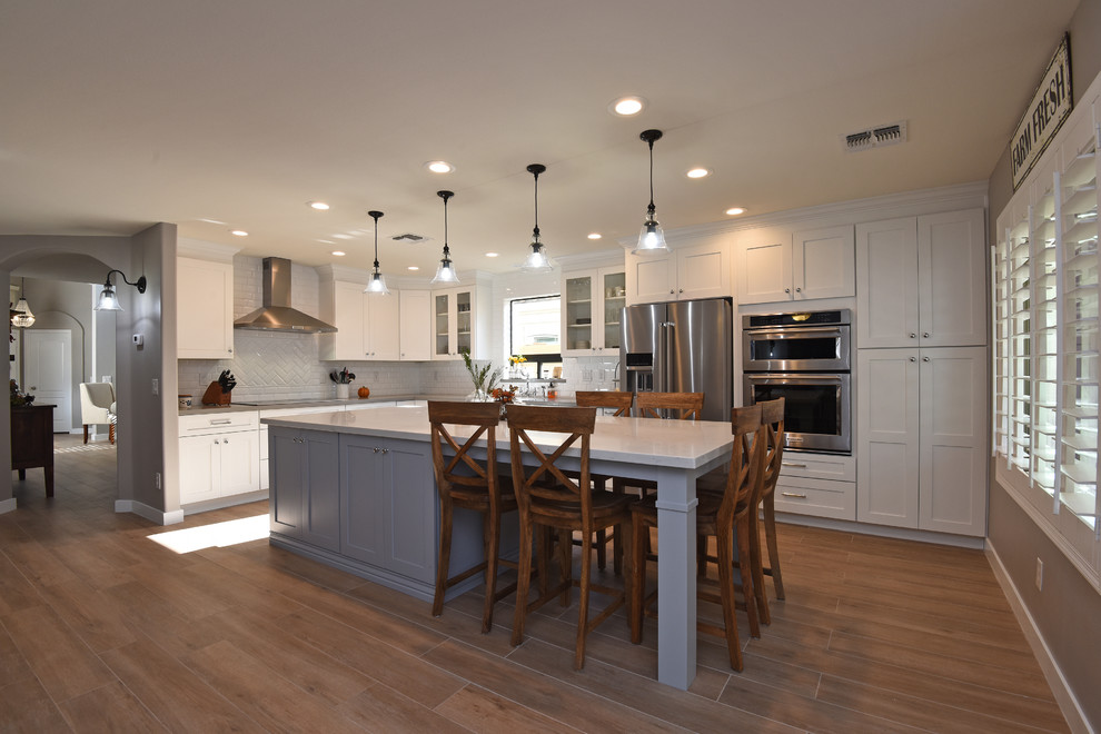 Foto på ett mellanstort lantligt kök och matrum, med en rustik diskho, skåp i shakerstil, vita skåp, vitt stänkskydd, rostfria vitvaror, mellanmörkt trägolv, en köksö och brunt golv
