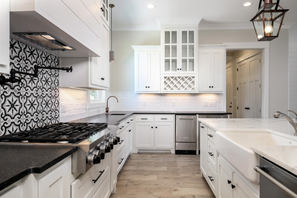 Example of a large beach style u-shaped light wood floor and brown floor eat-in kitchen design in New Orleans with a farmhouse sink, shaker cabinets, white cabinets, quartz countertops, white backsplash, subway tile backsplash, stainless steel appliances, an island and gray countertops