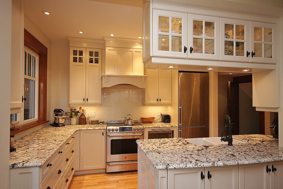 Example of a mid-sized transitional l-shaped light wood floor eat-in kitchen design in Ottawa with a farmhouse sink, shaker cabinets, white cabinets, granite countertops, white backsplash, subway tile backsplash, stainless steel appliances and an island