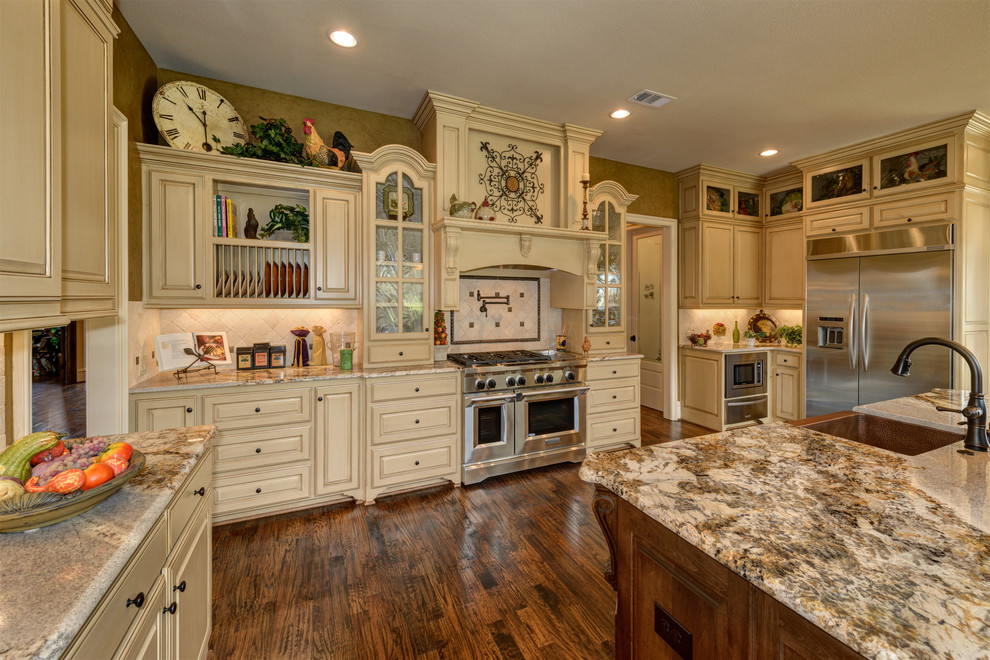 Large french country u-shaped dark wood floor and brown floor open concept kitchen photo in Dallas with a farmhouse sink, distressed cabinets, granite countertops, beige backsplash, stainless steel appliances, an island, stone tile backsplash and raised-panel cabinets