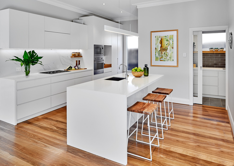 Example of a trendy galley medium tone wood floor and brown floor kitchen design in Sydney with an undermount sink, flat-panel cabinets, white cabinets, stainless steel appliances, an island, white countertops and white backsplash