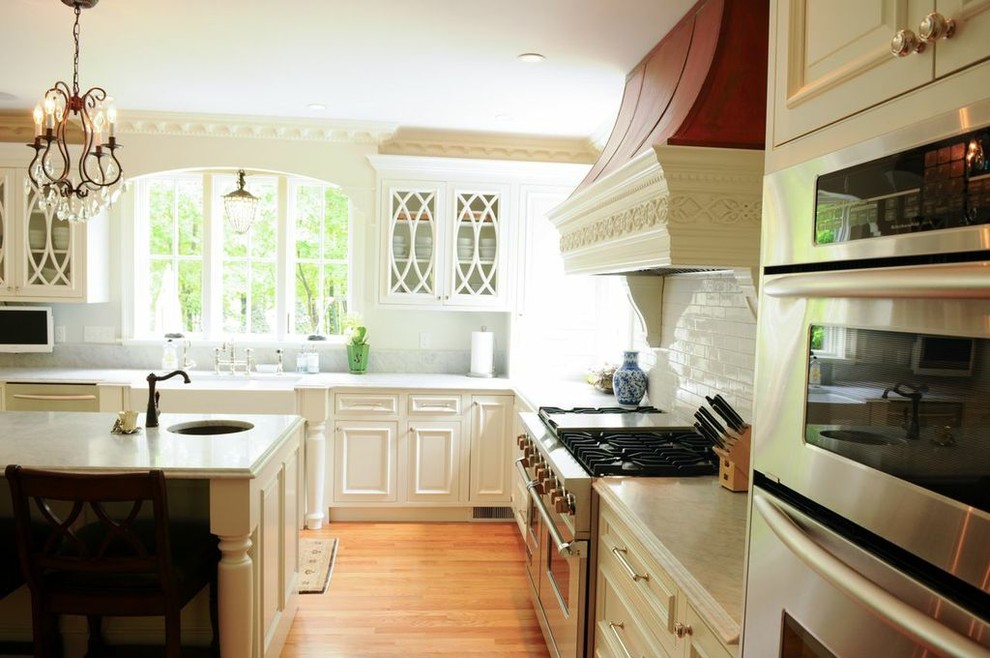 Large elegant light wood floor eat-in kitchen photo in Boston with a farmhouse sink, raised-panel cabinets, white cabinets, marble countertops, subway tile backsplash, stainless steel appliances, white backsplash and an island
