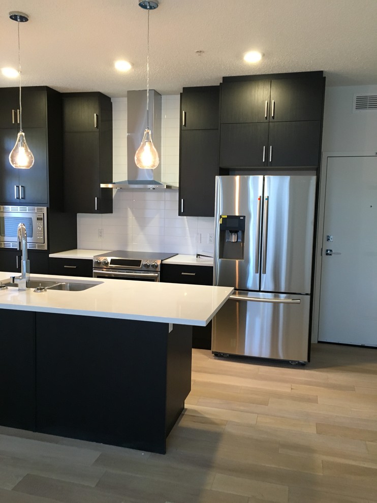 Medium sized modern galley kitchen/diner in Calgary with a submerged sink, flat-panel cabinets, black cabinets, white splashback, metro tiled splashback, stainless steel appliances, light hardwood flooring and an island.