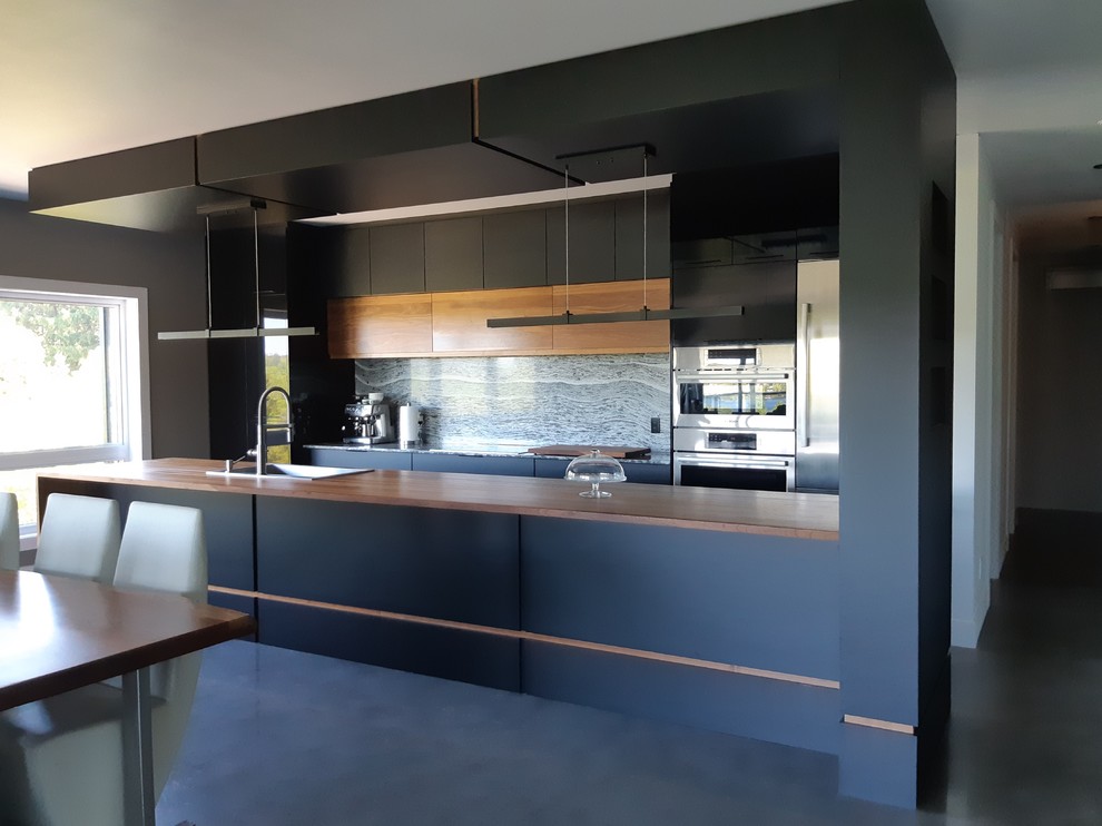 Large modern galley kitchen in Montreal with flat-panel cabinets, black cabinets, wood worktops, grey splashback, stone slab splashback, stainless steel appliances, concrete flooring, an island, grey floors and brown worktops.