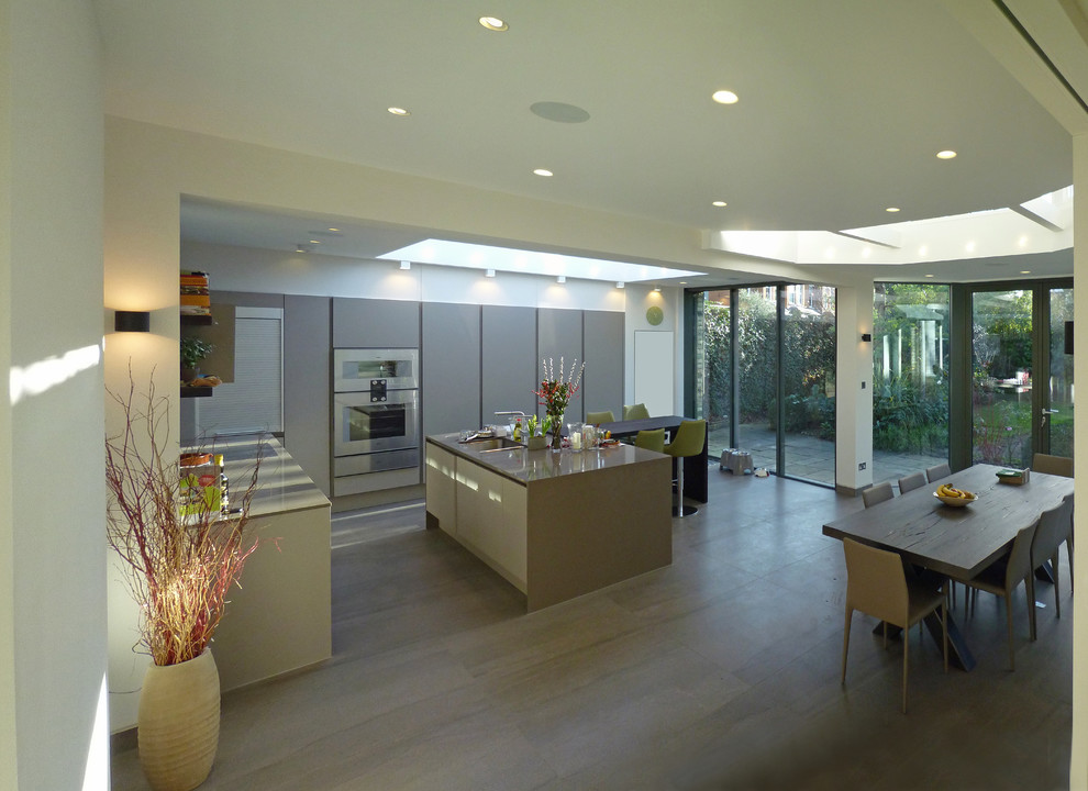 This is an example of a contemporary l-shaped kitchen/diner in London with an integrated sink, flat-panel cabinets, grey cabinets, composite countertops, metallic splashback, mirror splashback, stainless steel appliances, porcelain flooring and an island.