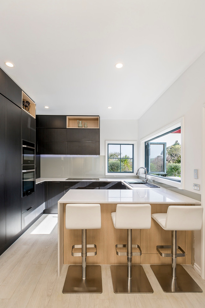 Mittelgroße Moderne Wohnküche in U-Form mit Einbauwaschbecken, flächenbündigen Schrankfronten, dunklen Holzschränken, Laminat-Arbeitsplatte, Küchenrückwand in Grau, Glasrückwand, Laminat, Halbinsel und weißer Arbeitsplatte in Auckland