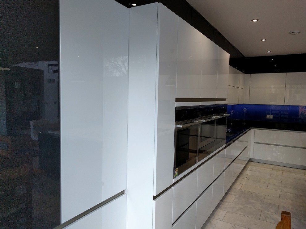 Bild på ett stort funkis kök, med en integrerad diskho, släta luckor, vita skåp, bänkskiva i kvartsit, blått stänkskydd, glaspanel som stänkskydd, svarta vitvaror, klinkergolv i keramik och en köksö